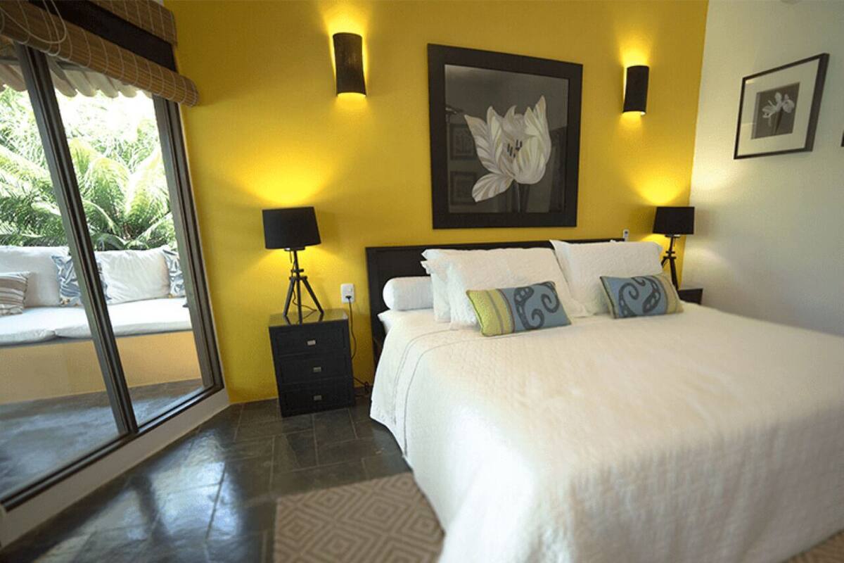 A bedroom with a large bed, two bedside tables, a window with a view of greenery, and a wall-mounted picture.