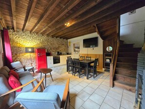 A cozy living room with a stone wall, wooden beams, a red refrigerator, a TV, a dining table with chairs, and a staircase.