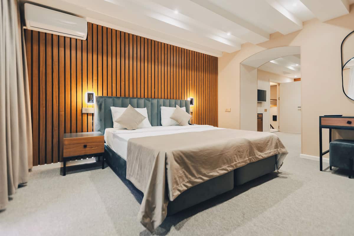 A modern hotel room with a wooden panel wall, a large bed with a headboard, bedside tables, a desk, and a mirror.