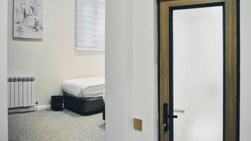 Quarto com cama, radiador, janela com persianas e porta com painel de vidro fosco.