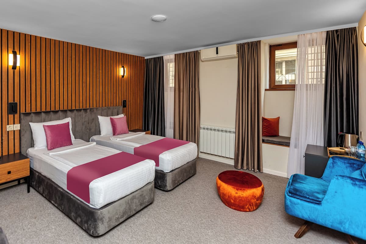 A hotel room with two beds, a blue armchair, a round ottoman, a wooden accent wall, and a view of the outside.