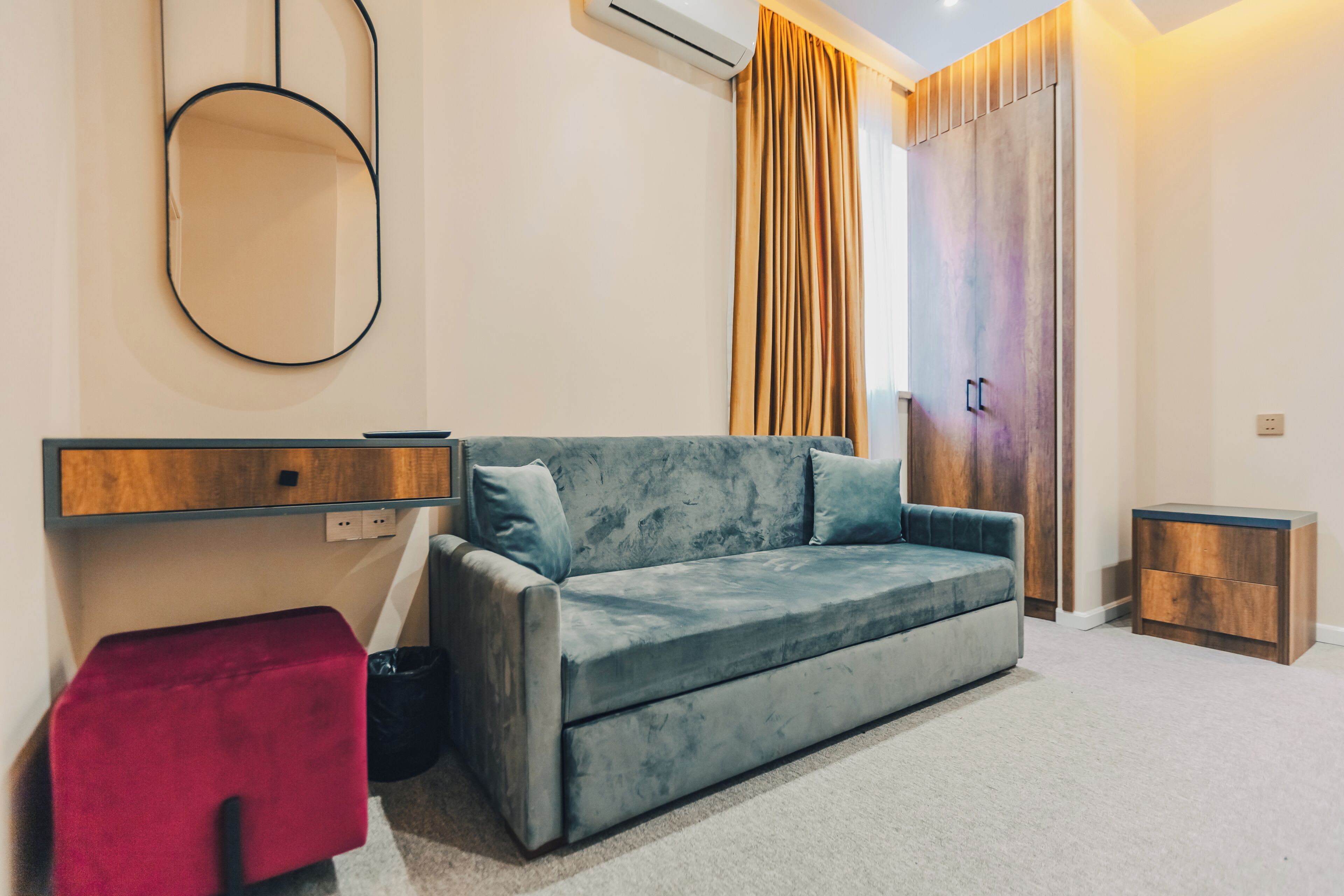 A modern room with a grey sofa, a red ottoman, a wooden side table, and a large mirror on the wall.