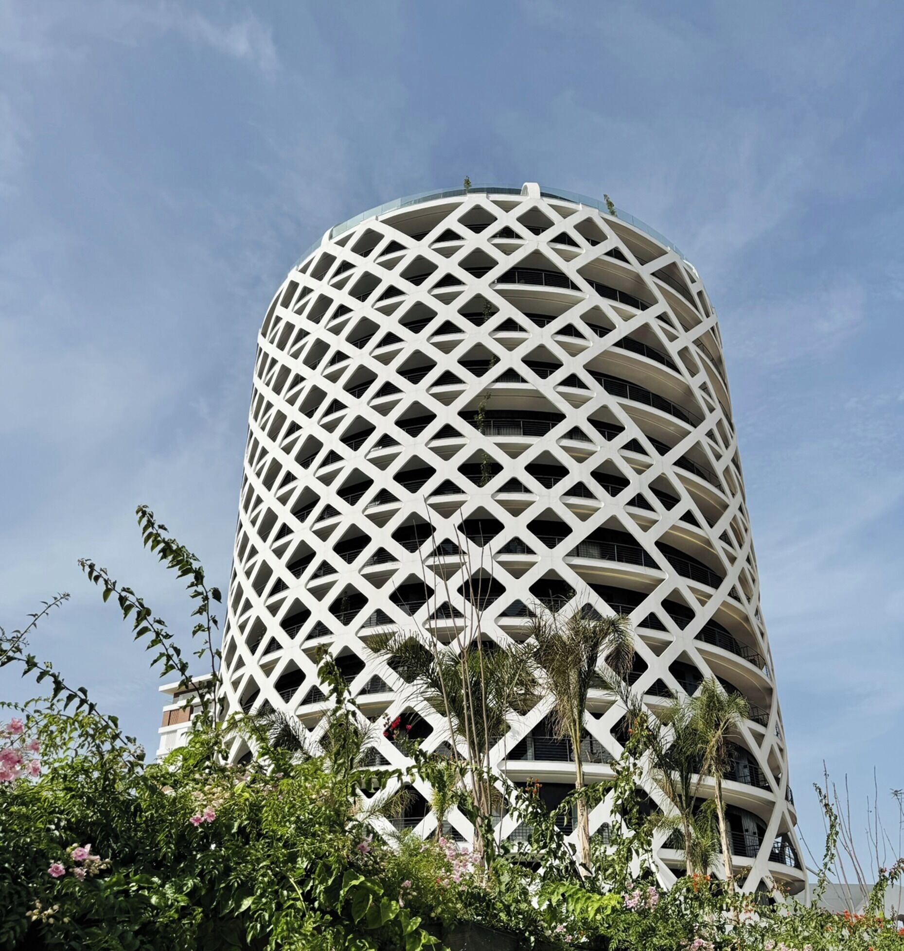 Un bâtiment cylindrique à la façade ajourée, entouré de verdure et de fleurs.