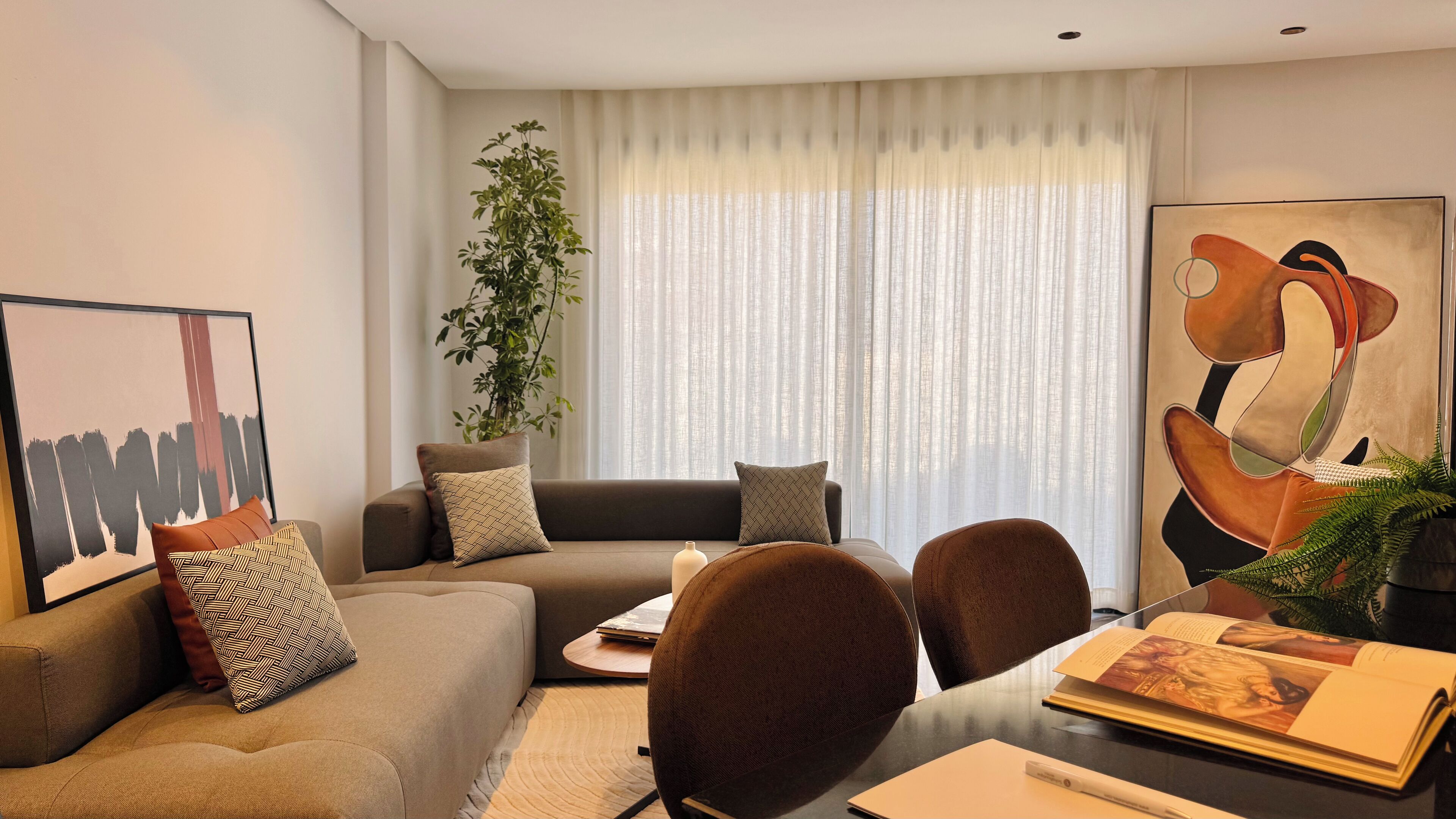 Un salon moderne comprenant un canapé, des fauteuils, une table basse et une table à manger. Une grande fenêtre est agrémentée de rideaux légers, et on y trouve également une plante en pot et une œuvre d’art encadrée accrochée au mur.