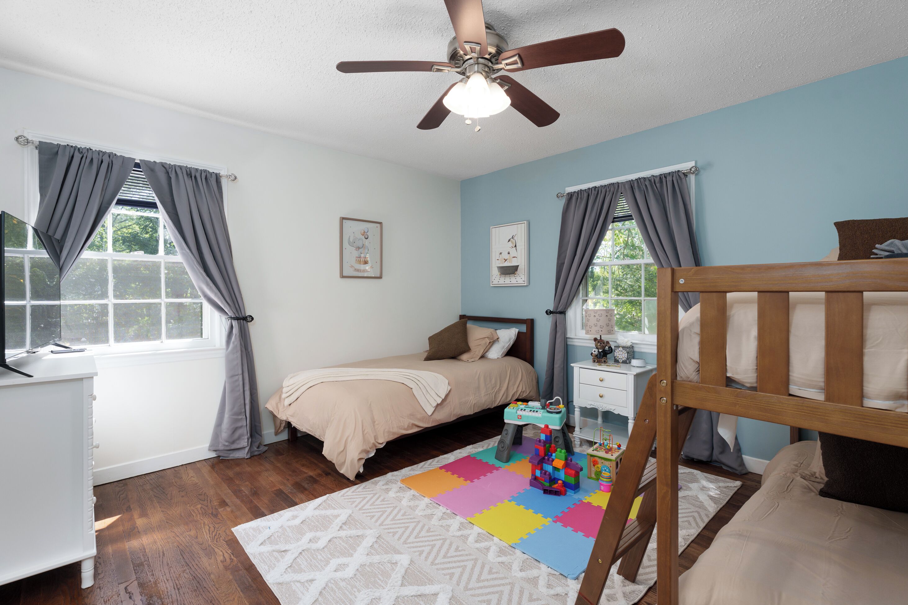 Une chambre à coucher comprenant un lit, un bureau, une chaise, un ventilateur de plafond et une fenêtre avec des rideaux.