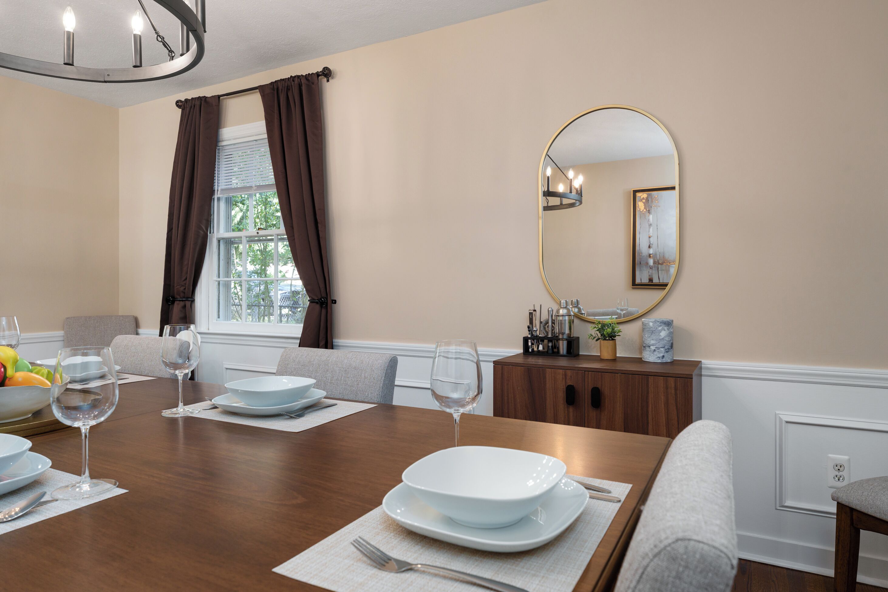 Un coin repas avec une table en bois mise pour quatre personnes, comprenant des assiettes, des verres et des couverts. Il y a un miroir au mur et une fenêtre avec des rideaux.