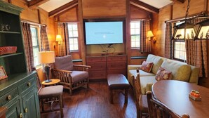 Een knus kamer in chaletstijl met een houten inrichting, een groot televisiescherm, een bank, een eettafel en een houten kast.