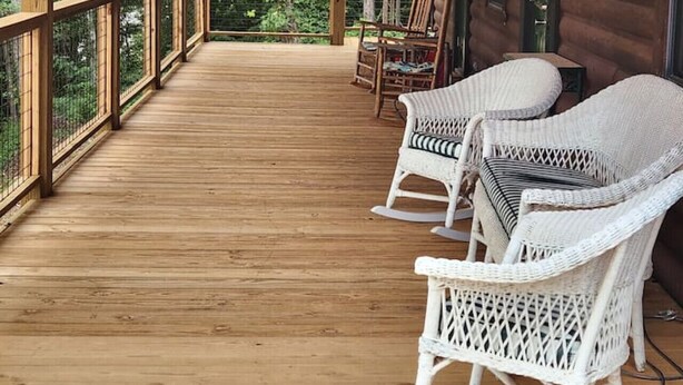 Een houten terras met wit rieten meubilair, waaronder stoelen en een tafel, met uitzicht op een bebost gebied.