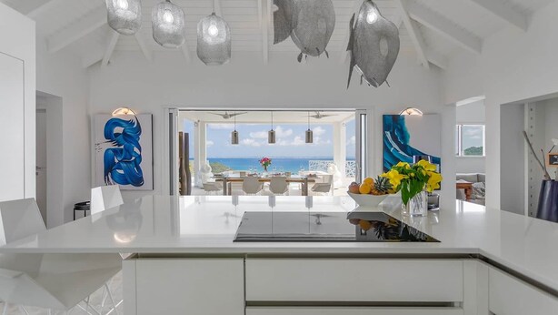 Eine moderne Küche mit einer großen Kochinsel, weißen Schränken und Pendelleuchten. Durch große Fenster ist der Blick aufs Meer möglich.