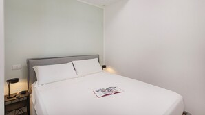 Una cama bien hecha con sábanas y almohadas blancas, una mesita de noche con una lámpara y un control remoto, y una revista sobre la cama.