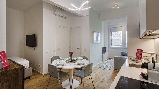 Una cocina moderna con una máquina Nespresso integrada, un comedor con una mesa redonda y sillas grises, y una pequeña zona de estar con un sofá gris.