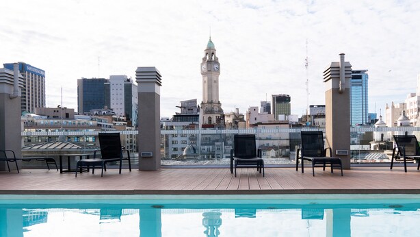 Una piscina en la azotea con sillones de descanso, vista a un paisaje urbano que incluye una torre del reloj y edificios modernos.