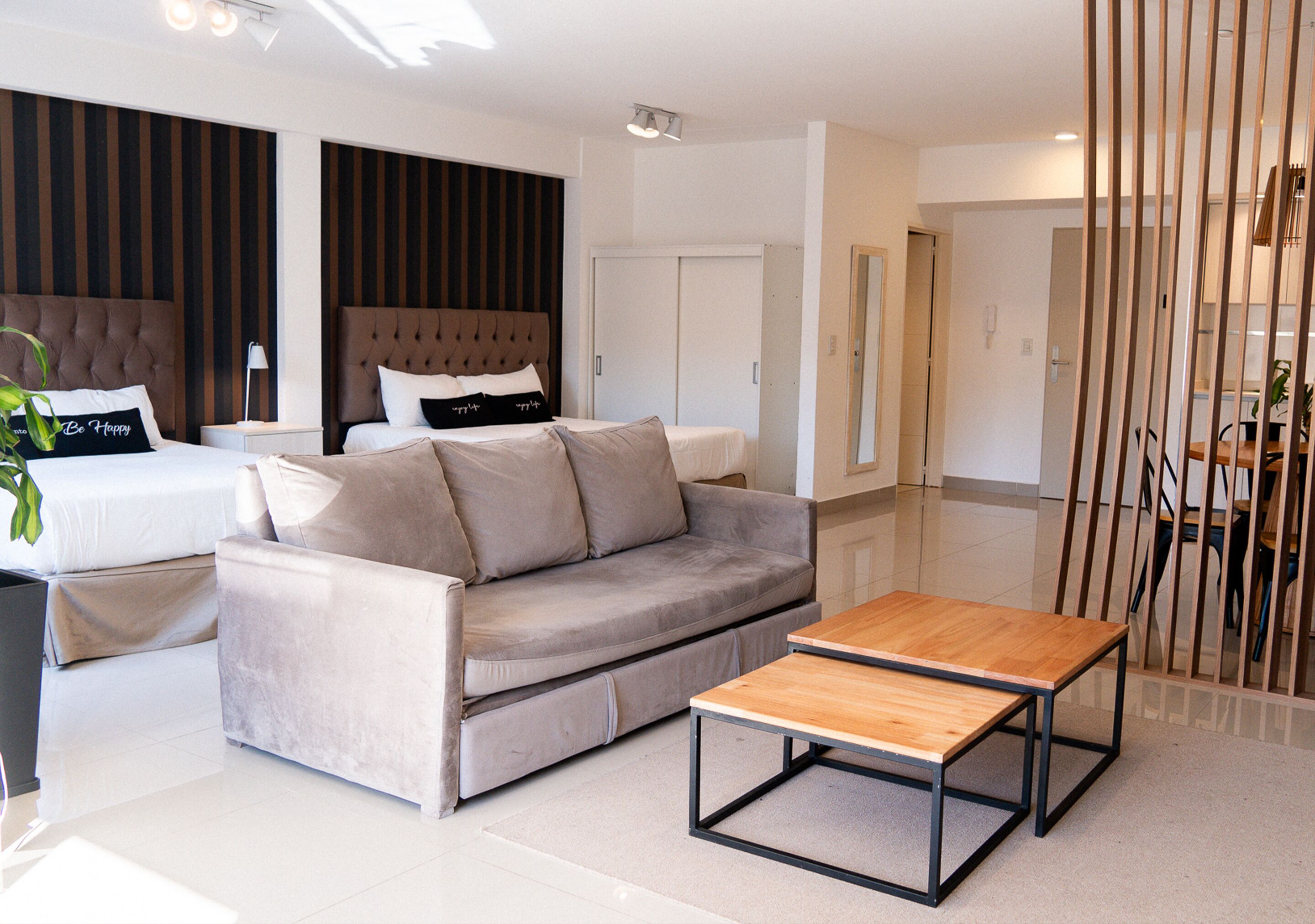 A modern hotel room with a bed, a sofa, a wooden coffee table, and a dining area with a table and chairs.