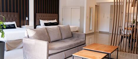 A modern hotel room with a bed, a sofa, a wooden coffee table, and a dining area with a table and chairs.