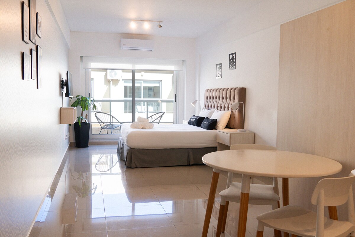 Habitación de hotel moderna con cama, una mesita y sillas. Tiene un balcón con vistas al exterior.