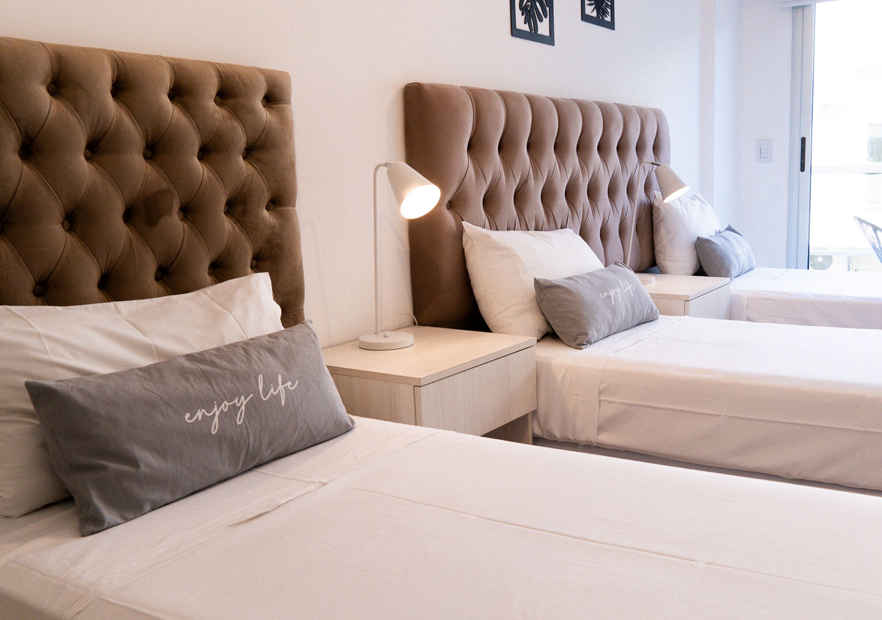 A hotel room with two beds, each with a tufted headboard, a bedside table with a lamp, and a pillow with the text "enjoy life".