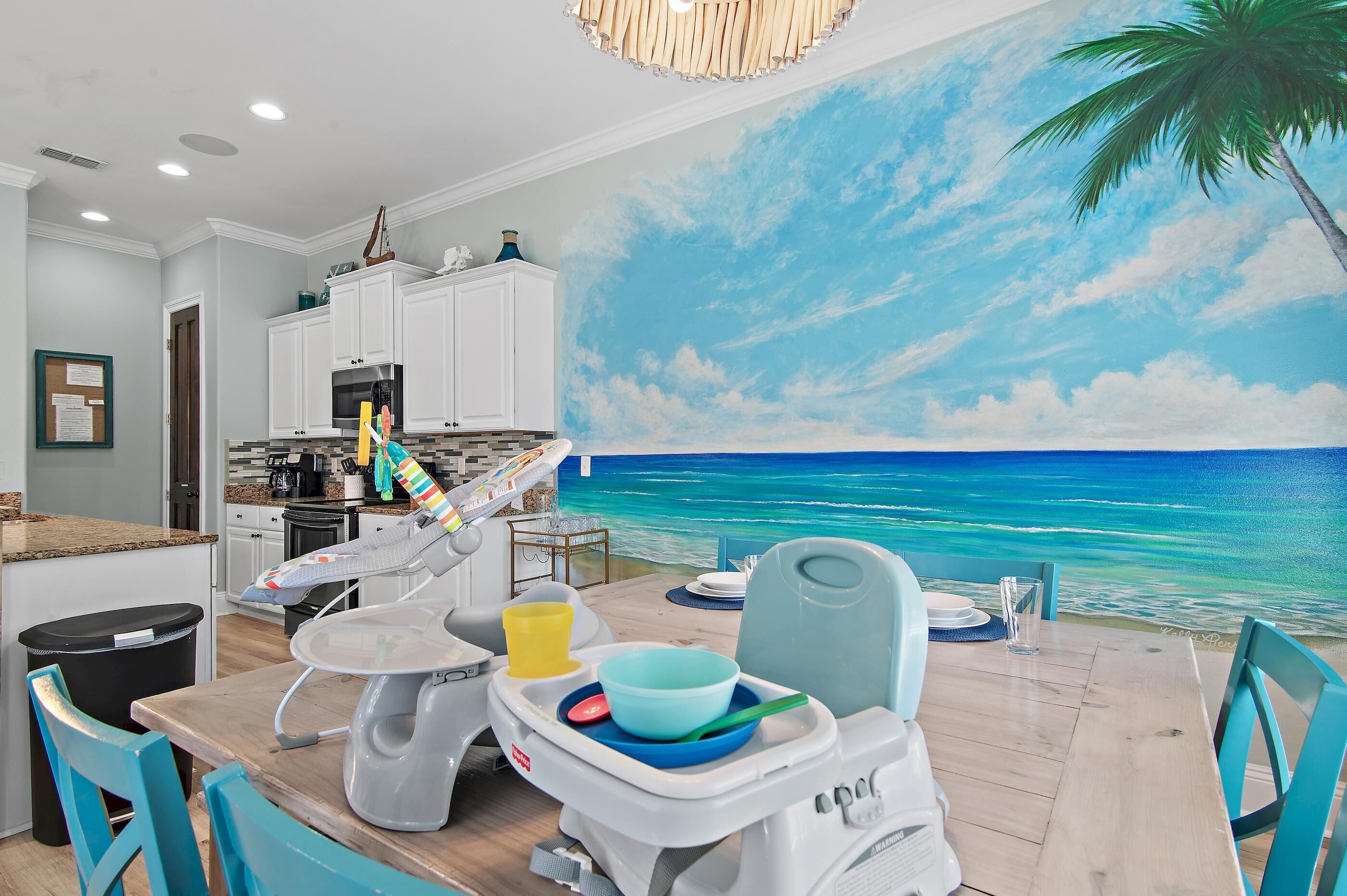A dining area with a view of the ocean, a kitchen with white cabinets, and a children's high chair.