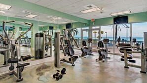 Une salle de sport équipée de divers appareils d’exercice, dotée d’un grand miroir et offrant une vue sur l’océan grâce à de larges fenêtres.