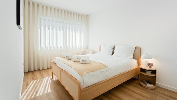 A modern hotel room with a wooden bed, white bedding, a bedside lamp, and a window with sheer curtains.