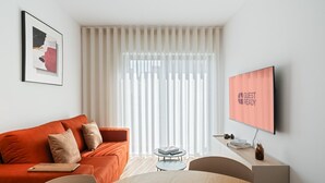 A modern hotel room with a large window, a flat-screen TV, a wooden table, and a red sofa.