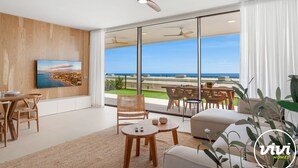Un salón moderno con un ventanal grande que da a vistas costeras, un ventilador de techo y un comedor con mobiliario de mimbre.