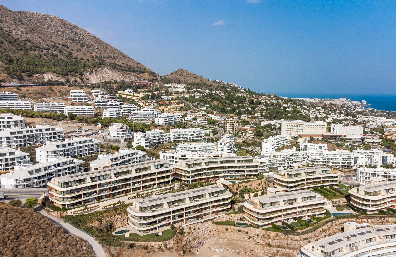 A coastal residential area with modern multi-story buildings, a clear view of the sea, and a hilly backdrop.