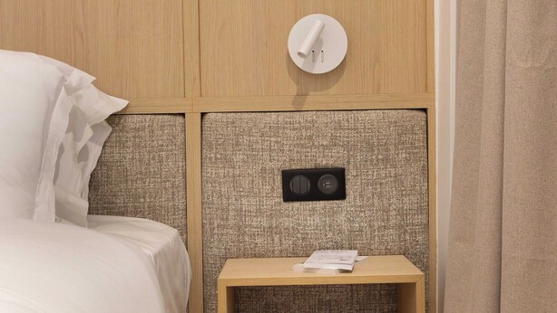 A neatly made bed with white linens, a small wooden nightstand with a lamp, and a textured headboard. - Beautiful Apartment- Jardin de Luxembourg (Paris)