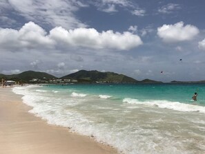 Une plage à l’eau turquoise et limpide, un rivage sablonneux et, au loin, des montagnes sous un ciel partiellement nuageux.