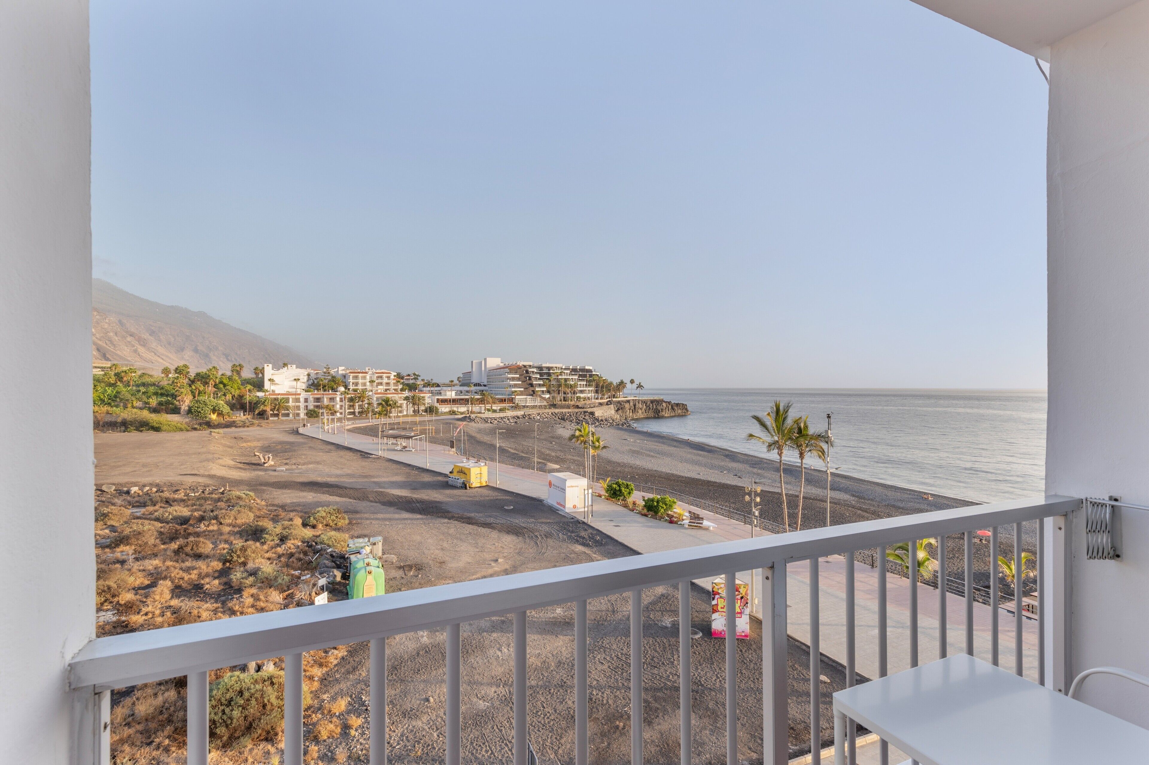 A balcony with a view of a beach, a building with multiple floors, and a clear sky.