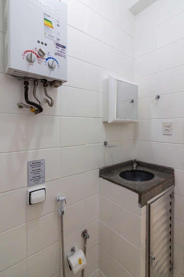 Un baño con inodoro, lavamanos y ducha. Hay un calentador de agua con panel de control, un secamanos y un dispensador de toallas de papel.