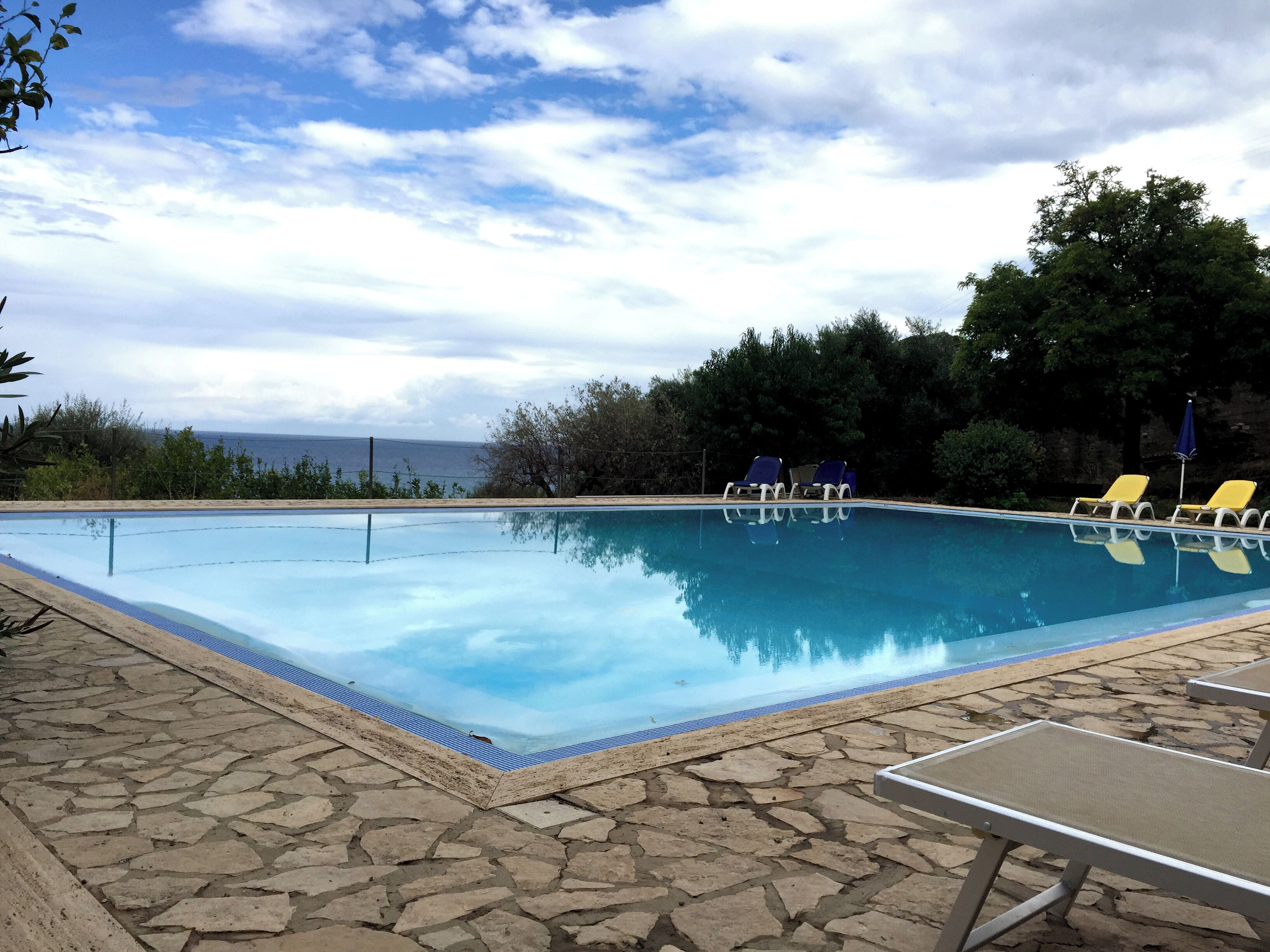 Piscina com deck de pedra, rodeada por árvores e vista para o mar.