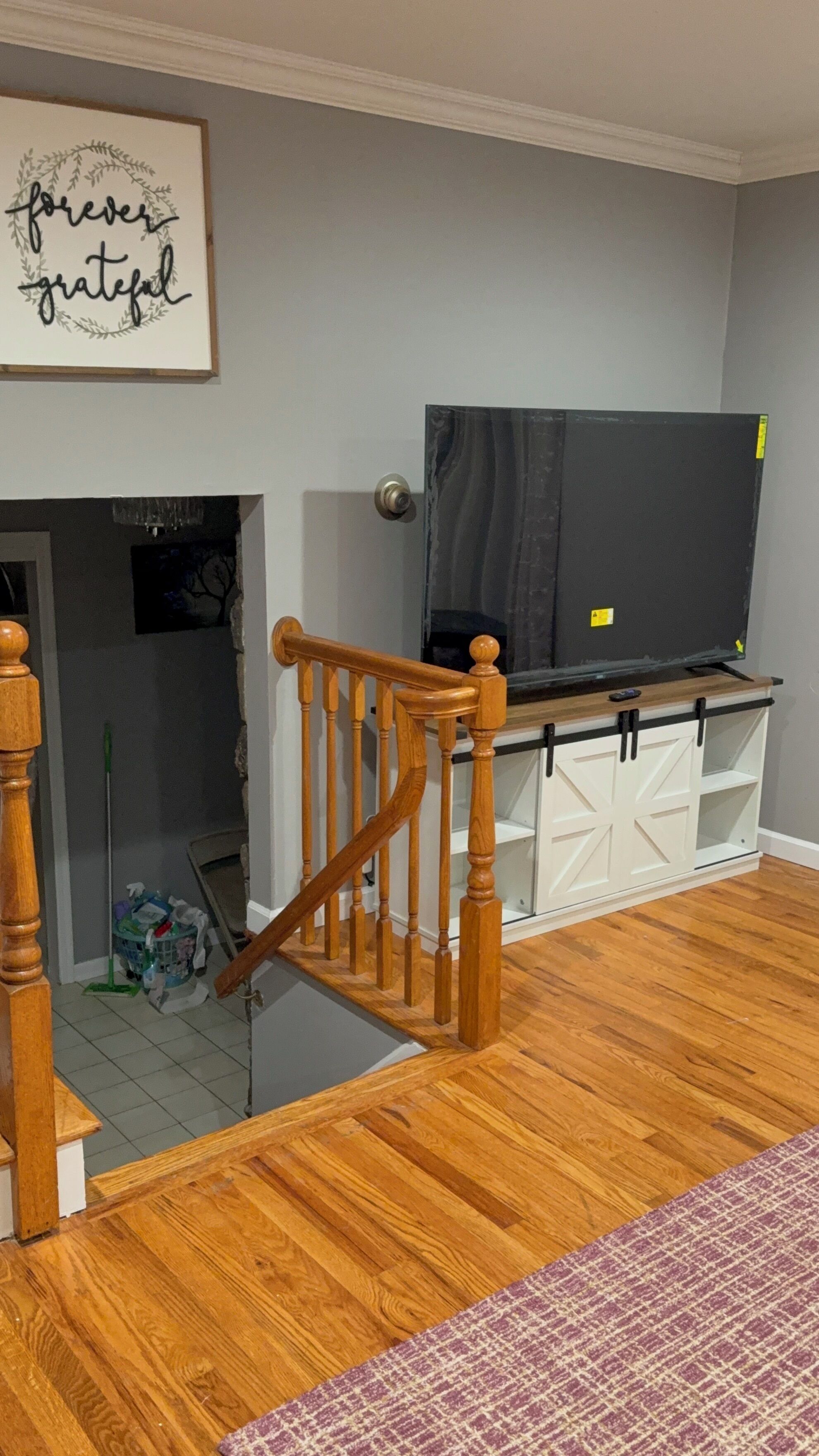 A staircase with wooden balusters leading to a landing. A flat-screen TV is mounted on a wooden shelf above a white cabinet with black handles.