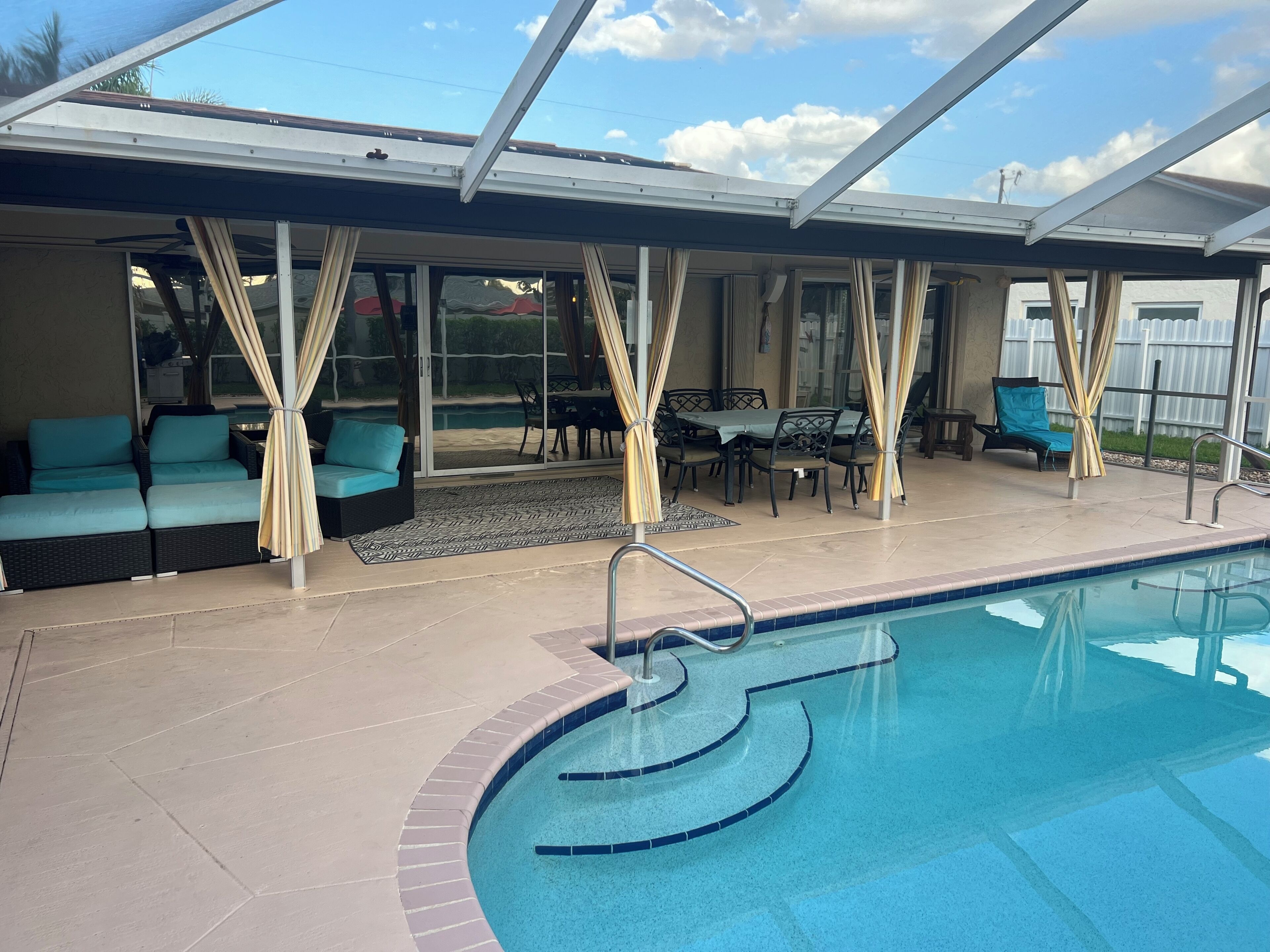 A swimming pool with a glass enclosure, a covered seating area with chairs and a table, and a view of the outdoors.