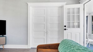 Una habitación con un sofá marrón, un televisor sobre un mueble bajo, una puerta blanca y una ventana con persianas.