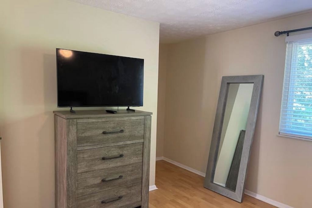 Una habitación con un aparador de madera, un televisor de pantalla plana encima y un espejo grande apoyado en la pared, además de una ventana con persianas.