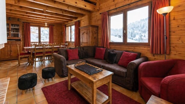 A wooden cabin interior with a wooden dining table, a sofa, a red armchair, and a lamp.