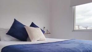A bedroom with a large bed, white and blue bedding, a bedside table with a lamp, and a window with a city view. - ✨ Spacious residence in Manta | Private pool, BBQ and privileged location (Manta)