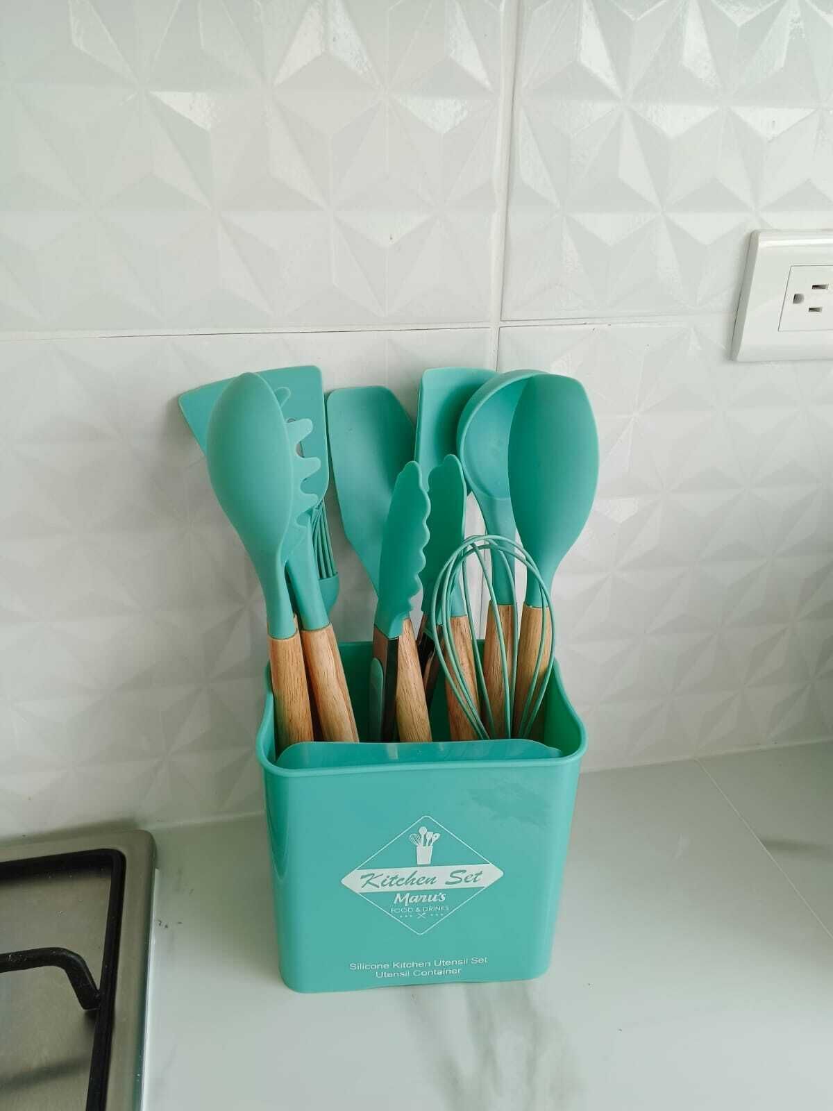 Un ensemble d’ustensiles de cuisine en silicone de couleur turquoise, présenté dans un récipient orné d’un logo, est posé sur un plan de travail.
