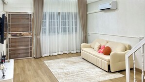 Un salon moderne avec un canapé beige, un lustre et une grande fenêtre ornée de rideaux légers.