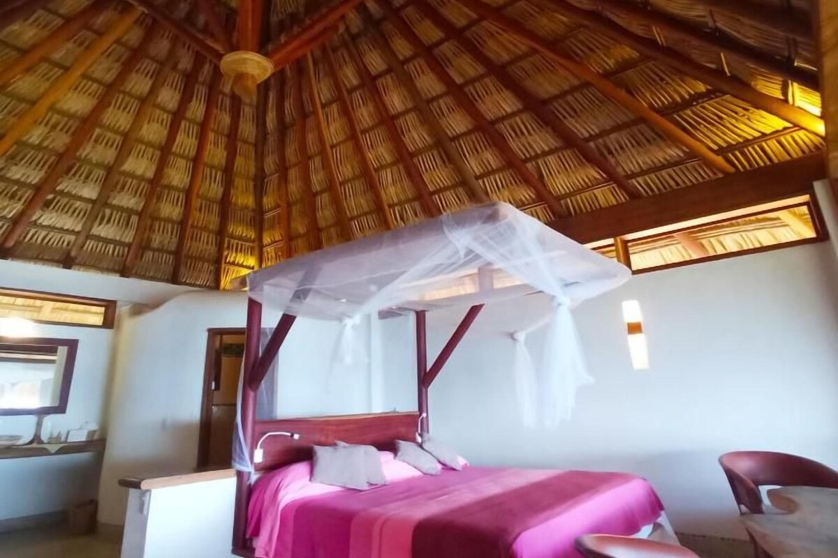 A room with a bed under a thatched roof, a bedside table, and a chair.