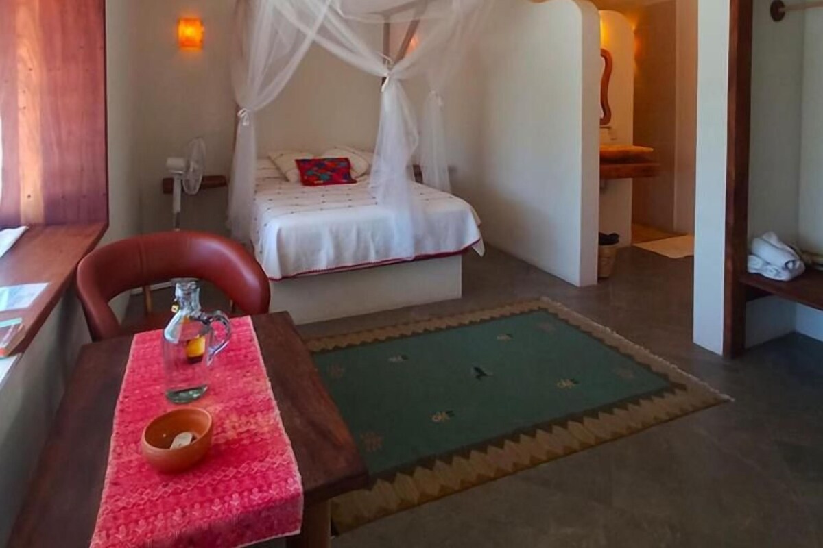 A room with a bed under a white canopy, a table with a red tablecloth, a ceiling fan, and a green rug.