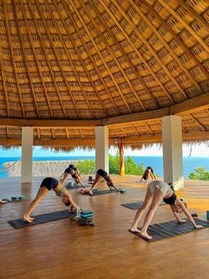 Un grupo de personas practicando yoga sobre esterillas bajo una estructura de techo de paja con vista al océano.