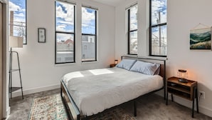 A bedroom with a bed, two bedside tables, a floor lamp, and large windows with a view of buildings and sky.