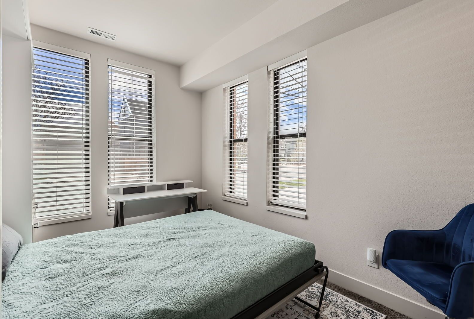 A bedroom with a bed, a desk, and a chair. There are windows with blinds.