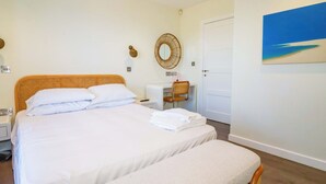 Una cama bien hecha con ropa blanca, una mesita de noche con una lámpara, una silla y un escritorio con otra silla. Hay una puerta y un cuadro en la pared.