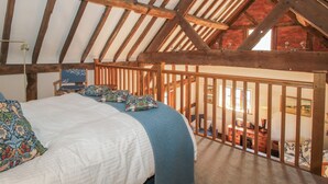 A bedroom with a sloped ceiling, wooden beams, a bed with patterned pillows, and a view of the living area below.