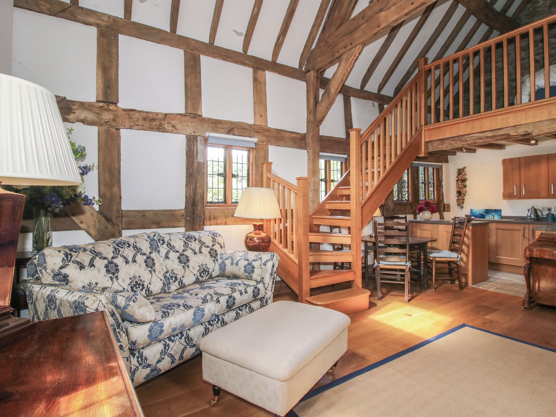 A rustic interior with exposed wooden beams, a wooden staircase, a floral sofa, and a dining area.