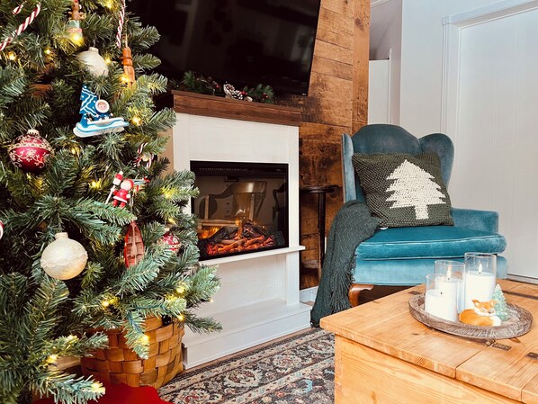 A cozy living room with a fireplace, a Christmas tree, a blue armchair, and a coffee table with candles.