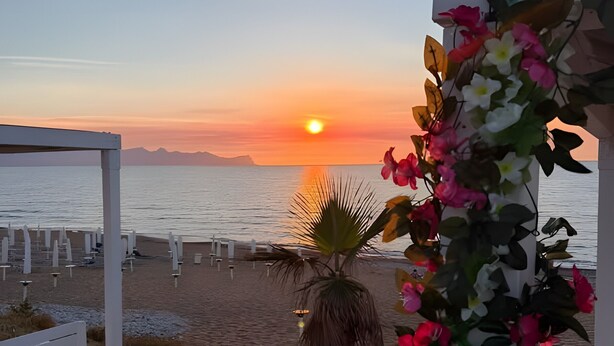 Een uitzicht op het strand met een zonsondergang, een palmboom en een decoratieve bloemboog.