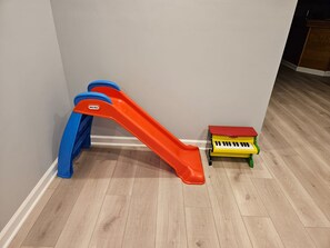 A room with a wooden floor, a red and blue slide, and a small yellow and green toy piano. - Fun 5BR 4.5BA, Pool, Hot Tub, Game Rm, Near O’Hare (Wood Dale)